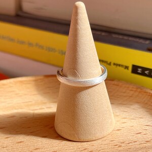 May include: A silver ring with a brushed finish sits on a wooden ring display stand.
