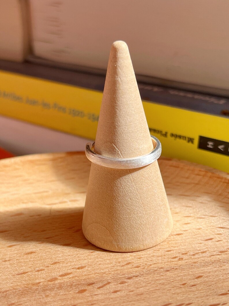 May include: A silver ring with a brushed finish sits on a wooden ring display stand.