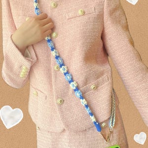 Puede incluir: Un conjunto de chaqueta y falda de tweed rosa con una correa de teléfono de flores azules y blancas. La correa está unida a una funda de teléfono verde.