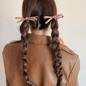 Könnte beinhalten: Zwei lange Zöpfe, die mit kleinen, hellbraunen Schleifen-Haarspangen gehalten werden. Die Person trägt eine braune Lederjacke. Die Zöpfe sind dick und sauber geflochten und ergeben eine klassische und elegante Frisur.