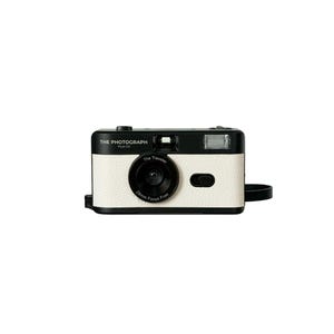 Puede incluir: Una cámara de estilo retro con cuerpo blanco y detalles negros. La cámara tiene una lente de 28 mm sin enfoque y el texto "The Photograph" está impreso en la parte superior. Una correa de muñeca negra está adjunta.