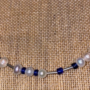 May include: A silver necklace with white pearls and blue glass beads. The necklace is on a brown fabric background.