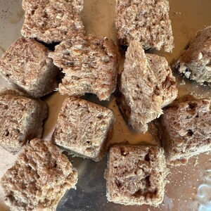 May include: A collection of roughly cut, square-shaped, light brown food items. The texture appears porous and fibrous, with a slightly uneven surface. The items are arranged on a metallic surface, suggesting a culinary context.