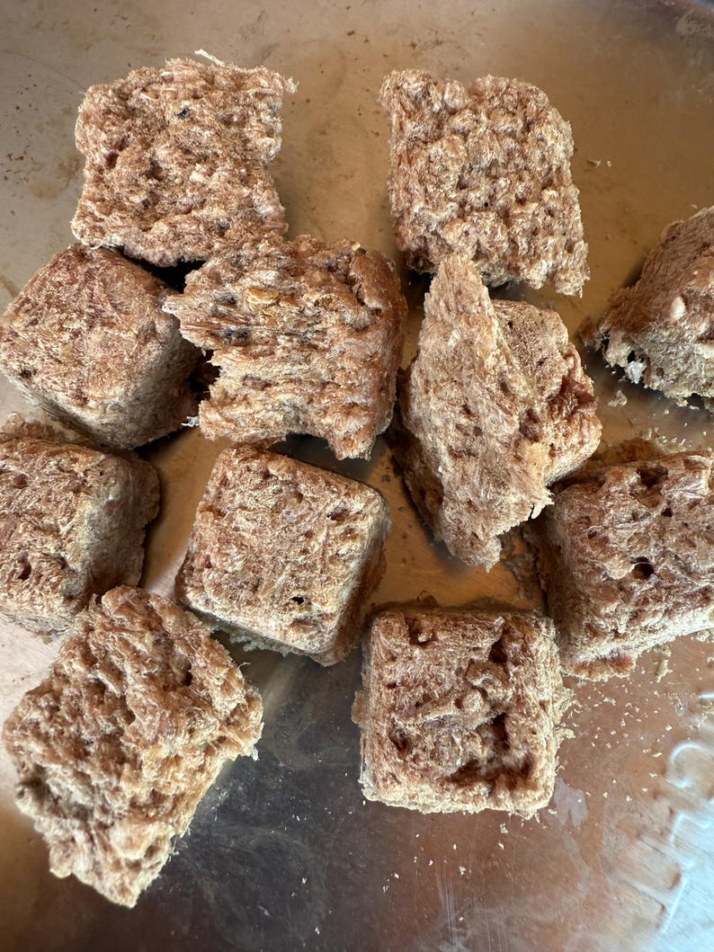 May include: A collection of roughly cut, square-shaped, light brown food items. The texture appears porous and fibrous, with a slightly uneven surface. The items are arranged on a metallic surface, suggesting a culinary context.