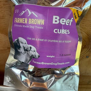 May include: A sealed silver bag of Farmer Brown Beef Cubes dog treats. The purple label features the brand name, a mountain graphic, and the text "Colorado Made Dog Treats." The bag contains 1.4 ounces of freeze-dried beef cubes.