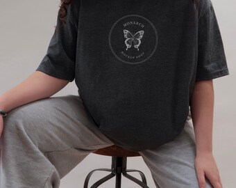 May include: Dark gray t-shirt with a circular graphic featuring a butterfly and the words "MONARCH MOCKUP SHOP" in white. The shirt is a crew neck and has short sleeves. The model is wearing gray sweatpants.
