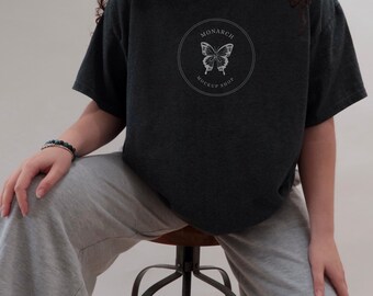 May include: Dark gray t-shirt with a circular graphic featuring a butterfly and the text "MONARCH MOCKUP SHOP". The shirt is worn with gray sweatpants. A person is sitting on a brown stool.