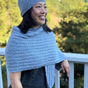 May include: A light blue knitted hat and matching shawl. The hat is a close-fitting beanie style. The shawl is draped over the shoulders and has a textured appearance. The outfit is made of a soft, possibly wool, material.