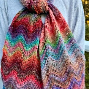 May include: A colorful, hand-knitted scarf with a chevron pattern. The scarf features a vibrant mix of colors, including red, orange, yellow, green, blue, and purple. The scarf is made of soft yarn and is perfect for adding a pop of color to any outfit.