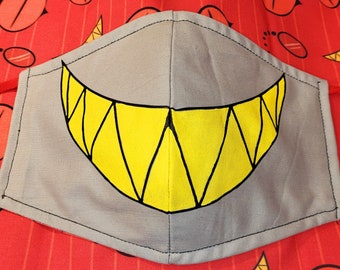 May include: A gray fabric face mask with a yellow and black smiling mouth design. The mouth is drawn with sharp teeth.