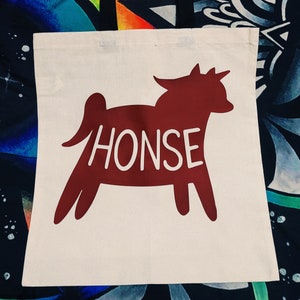 May include: A white canvas tote bag with a red silhouette of a horse with the word "HONSE" written in white across the body of the horse.