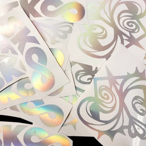 May include: A collection of iridescent vinyl stickers featuring various designs, including an infinity symbol, a swirl pattern, and a geometric shape. The stickers are on a white background.