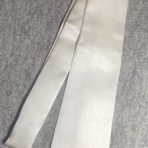 May include: Ivory-colored satin tie, laid diagonally on a gray surface. The tie has a smooth, lustrous texture and a classic pointed end. A formal accessory suitable for various occasions.