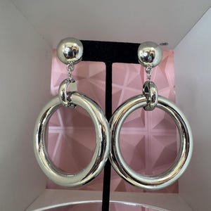 May include: A pair of silver-toned earrings featuring a polished spherical stud top connected by a small link to a large, thick circular hoop pendant. The metal surface is smooth and reflective, creating a high-shine finish on the circular hoops.