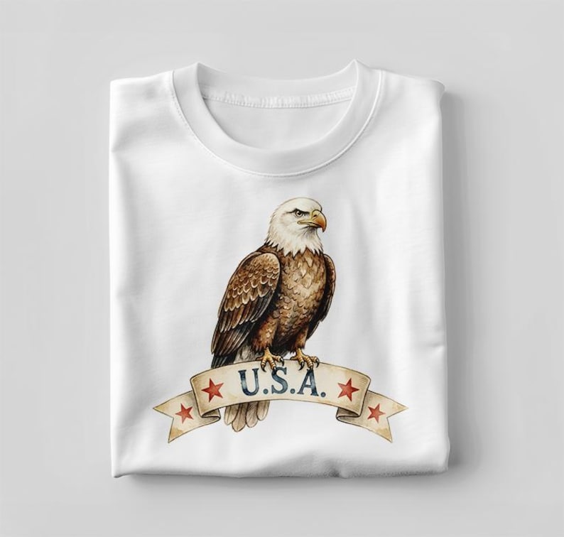 May include: A white crew neck t-shirt featuring a graphic of a bald eagle perched on a beige ribbon banner. The banner displays the text U.S.A. in dark blue lettering, accented by four small red stars positioned on the folded ends of the ribbon.