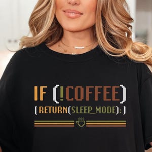 Op de afbeelding: Zwart T-shirt met een grafische print die "IF(!COFFEE) [RETURN(SLEEP_MODE)]" luidt in een retro lettertype. De grafiek is in tinten oranje, geel en bruin.