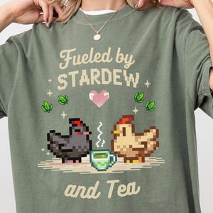 A woman wearing a green t-shirt with a pixelated design of two chickens and a cup of tea, along with the text "Fuelled by Stardew and Tea".