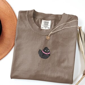 May include: A brown Comfort Colors t-shirt featuring a black cartoon chicken wearing a pink collar. A gold necklace with a pendant rests on the shirt. A brown hat and dried plant are in the background.