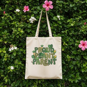 Peut inclure: Un sac fourre-tout crème avec l'inscription "PROUD PLANT PARENT" en vert, orné d'illustrations de plantes. Le sac, suspendu devant un fond de feuillage vert et de fleurs roses et blanches, est doté de longues anses.