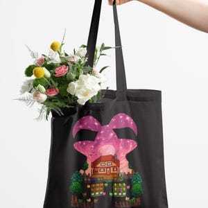 May include: Black tote bag with a pink and purple graphic of a house and trees. The bag is filled with a bouquet of flowers. The tote bag has long black handles.
