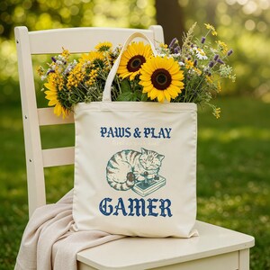 Op de afbeelding: Crèmekleurige draagtas met de tekst "PAWS & PLAY" en "GAMER" in blauw. De tas toont een slapende kat met een controller. Gevuld met zonnebloemen en andere bloemen, rustend op een witte stoel.