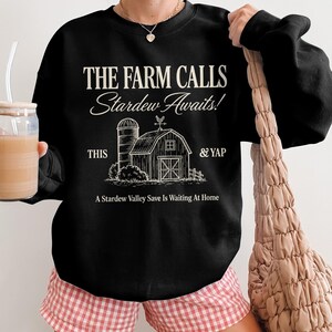 Könnte beinhalten: Schwarzes Sweatshirt mit dem Text "THE FARM CALLS Stardew Awaits!" und einer Grafik einer Scheune und eines Silos. Der Text "THIS & YAP" ist ebenfalls enthalten. Der Satz "A Stardew Valley Save Is Waiting At Home" ist darunter gedruckt.