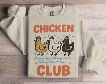 Może przedstawiać: Kremowa bluza z napisem "CHICKEN CLUB" w kolorze pomarańczowym. Poniżej tekstu znajdują się trzy kreskówkowe kurczaki w kolorze żółtym, białym i czarnym. Na koszulce znajduje się również napis "laying eggs, losing sleep, living the dream".