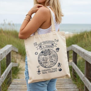 May include: Cream-colored tote bag with a whimsical design. The bag features the text "MY THERAPIST IS A JUNIMO" above a circular illustration of a village scene. Below, it reads "CHEAPER THAN REAL THERAPY".
