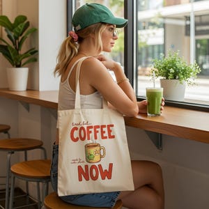 Peut inclure: Sac fourre-tout crème avec l'inscription "tired, but... COFFEE NOW" en rouge et une tasse à café dessinée avec une grenouille verte et une étoile. Le sac est porté par une personne portant une casquette verte et des lunettes.