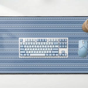 Op de afbeelding: Blauw en wit patroon bureaumat met een toetsenbord, muis en een glas ijskoffie. Het toetsenbord en de muis zijn lichtblauw. De bureaumat heeft een geometrisch ontwerp.