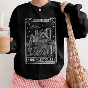 Könnte beinhalten: Schwarzes Sweatshirt mit weißer Grafik einer Farm, einschließlich Haus, Silo und Garten. Die Texte "LIFE ON THE FARM" und "THE VALLEY CALLS" sind weiß. Das Sweatshirt wird mit rosa-weiß karierten Shorts getragen.