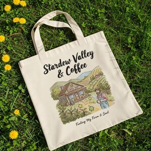 May include: A cream-coloured tote bag featuring the text "Stardew Valley & Coffee" and an illustration of a coffee shop in a rural setting. The bag also has the phrase "Fueling My Farm & Soul".