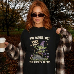 May include: A black crewneck t-shirt featuring a graphic of a green goblin wearing a purple wizard hat and cape, holding a sign that says NO. The text above reads THE OLDER I GET and below reads THE FUCKER THE NO, surrounded by stars and flowers.