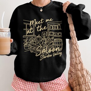 Könnte beinhalten: Schwarzes Sweatshirt mit einer goldenen Illustration mit dem Text "Meet me at the Saloon Stardew Valley". Das Design umfasst eine Holzkiste mit Produkten, ein Fass und Regale mit Gläsern. Die Person trägt eine rosa-weiß karierte Shorts.