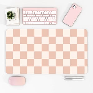 Puede incluir: Una alfombrilla de escritorio a cuadros rosa y crema con un teclado, un ratón y un teléfono rosa a juego. La alfombrilla de escritorio es rectangular y presenta un patrón repetido de cuadrados rosas y crema. Una pequeña planta suculenta en una maceta blanca.