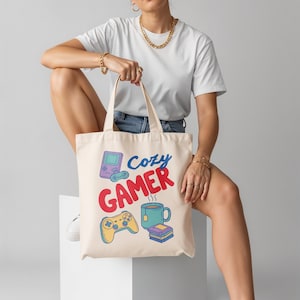 以下が含まれることがあります： クリーム色のトートバッグ。青と赤で「Cozy Gamer」の文字と、携帯ゲーム機、コントローラー、ティーカップ、本のイラストが描かれています。バッグには2つのハンドルが付いています。