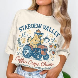 May include: Cream-colored t-shirt featuring a cartoon chicken driving a tractor, wearing sunglasses, and holding a coffee cup. The text "Stardew Valley" and "Coffee. Crops. Chaos." are printed above and below the illustration.
