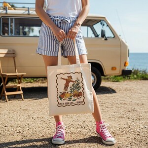 May include: Cream tote bag with a fall harvest design, including a scarecrow, pumpkins, and corn. The text "IT'S A GOOD LIFE... I GUESS" is printed below the illustration. The bag is held in front of a vintage van.