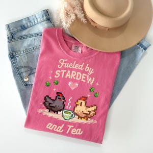 A pink t-shirt with a pixelated design of two chickens and a cup of tea, along with the text "Fuelled by Stardew and Tea" printed on it. The t-shirt is placed on top of a pair of jeans and a beige hat.