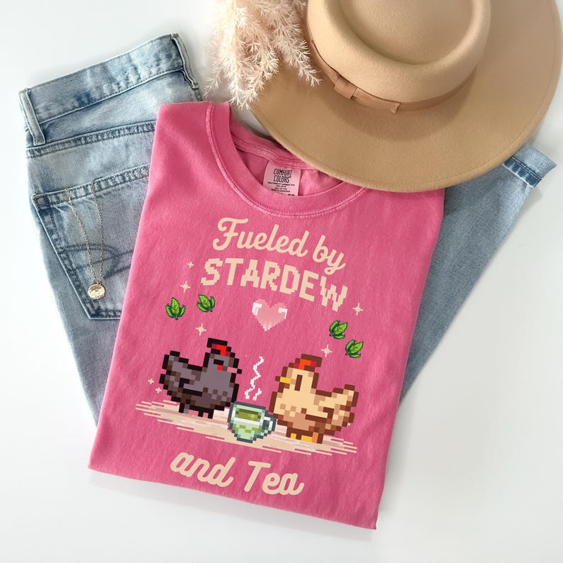 A pink t-shirt with a pixelated design of two chickens and a cup of tea, along with the text "Fuelled by Stardew and Tea" printed on it. The t-shirt is placed on top of a pair of jeans and a beige hat.