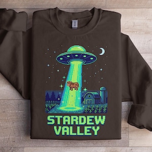 Könnte beinhalten: Ein braunes Sweatshirt mit einem pixeligen Design eines UFOs, das eine Kuh über einer Farm-Szene entführt. Der Text "STARDEW VALLEY" wird in grünen und blauen Pixelbuchstaben angezeigt.
