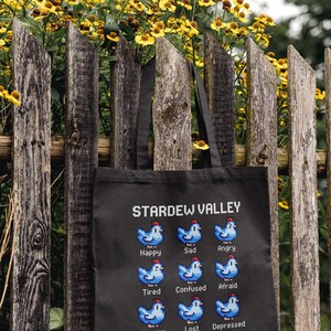 以下が含まれることがあります： 「Stardew Valley」の文字と、様々な感情を表すピクセル化された鶏が描かれた黒いトートバッグ。バッグは、背景に黄色の花がある、風化した木製のフェンスに掛けられています。