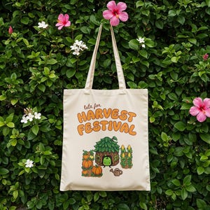 May include: A cream tote bag with the words "late for HARVEST FESTIVAL" in orange. The bag features a cartoon illustration of a house, pumpkins, corn, and a watering can. The bag has two cream handles and is set against a green leafy background.