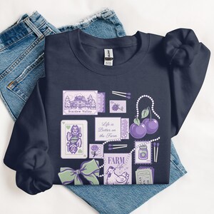 Könnte beinhalten: Marineblaues Sweatshirt mit einer Collage aus lila und weißen Illustrationen. Das Design umfasst ein Haus, Spielkarten, Kirschen und den Text "Life is Better on the Farm" und "Stardew Valley". Das Sweatshirt ist auf einer Jeans gefaltet.