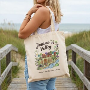 May include: A cream-coloured tote bag with a colourful illustration of a valley scene, including buildings, crops, and a river. The text "Junimo Valley" is above the illustration, and "Farming is Hard, Befriending is Harder" is below.