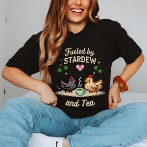A woman wearing a black t-shirt with a pixelated design of a chicken and a cup of tea, along with the text "Fuelled by STARDEW and Tea".