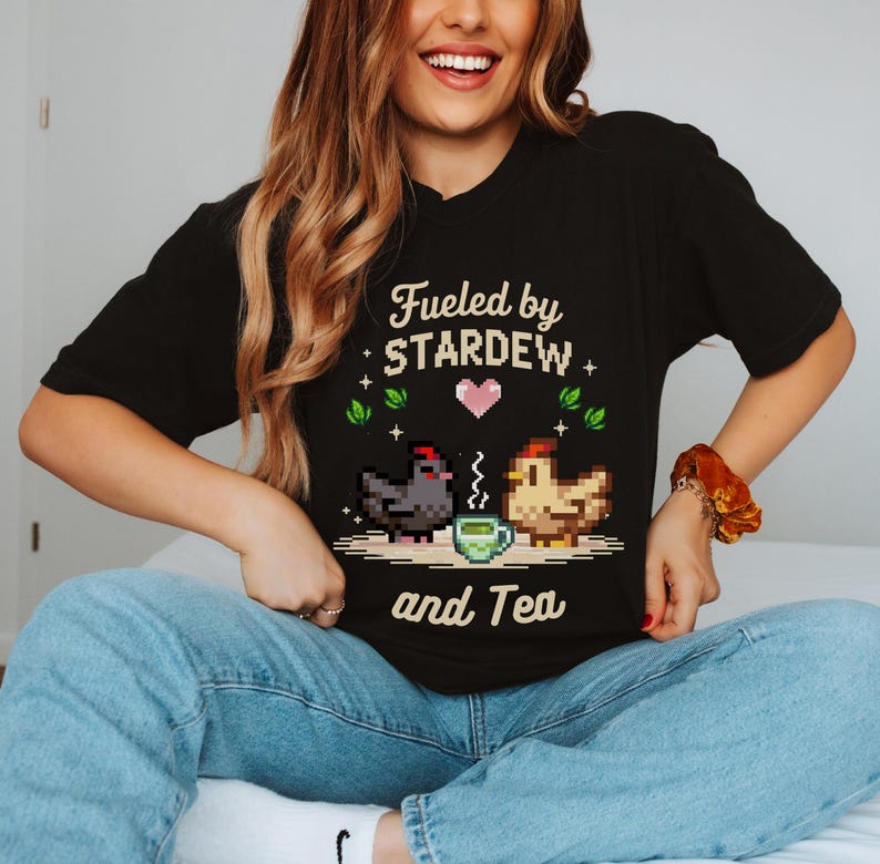 A woman wearing a black t-shirt with a pixelated design of a chicken and a cup of tea, along with the text "Fuelled by STARDEW and Tea".