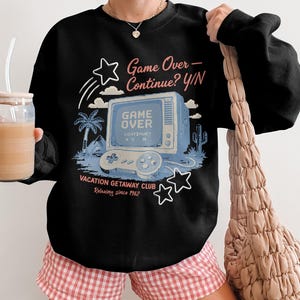 May include: Black sweatshirt with a retro video game graphic. The design features a vintage television screen with the words "Game Over" and a controller. The text "Vacation Getaway Club" and "Relaxing since 1962" are also included.