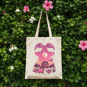 May include: A cream tote bag with a fantasy-themed design. The graphic shows a pink and purple silhouette of a creature with a starry sky, a house, and garden elements. The bag has two cream-colored handles, perfect for carrying items.