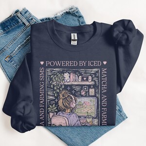 May include: Navy blue sweatshirt with a graphic featuring a person gaming, a computer, and the text "POWERED BY ICED MATCHA AND FARMING SIMS." The design includes a shelf with plants and books. The sweatshirt is laid on top of a pair of blue jeans.